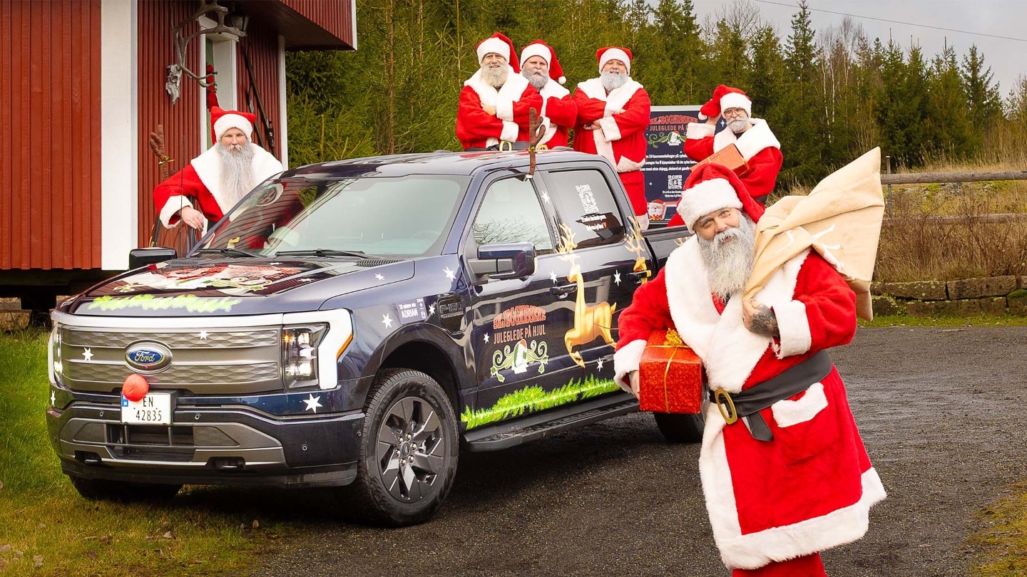 En nisse bærer en sekk med gaver. Resten av nissene står rundt F-150 Lightning.