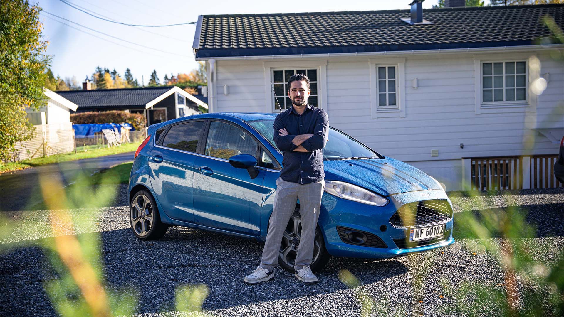 En person står med armene i kors foran en blå Ford Fiesta parkert på en grusvei.