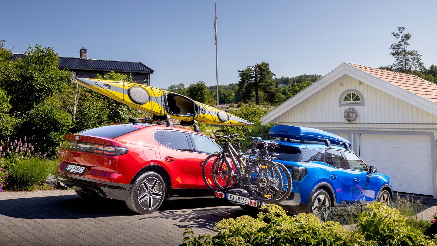 Rød helelektrisk Ford Capri med kajakk på taket og blå helelektrisk Ford Explorer med takboks og sykler på hengerfestestativ. 