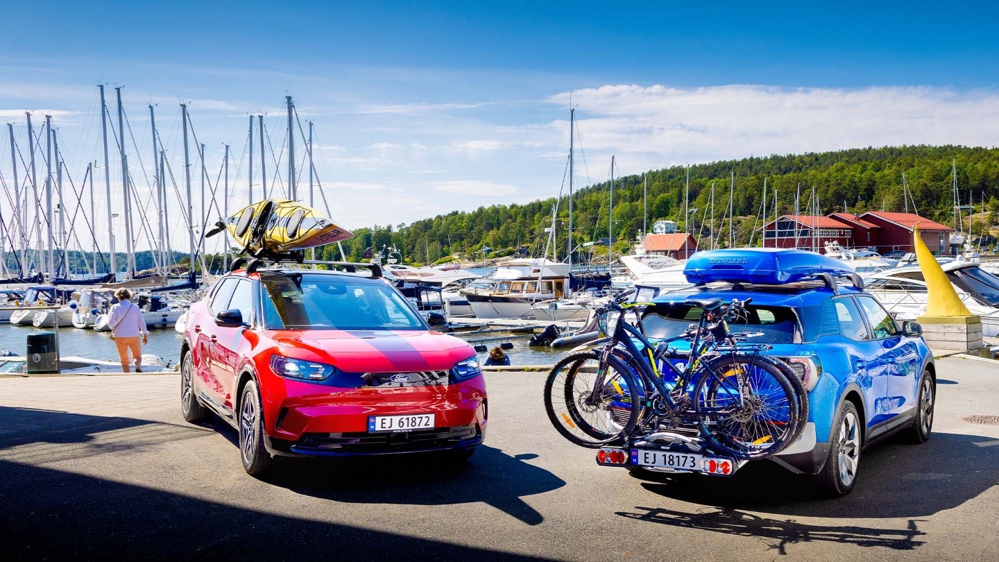 Rød helelektrisk Ford Capri med kajakk på taket og blå helelektrisk Ford Explorer med sykler på hengerfestestativ. 