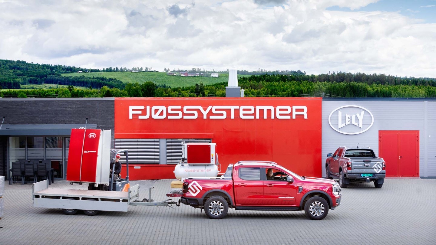 Rød Ford Ranger trekker en tilhenger med maskineri, parkert foran en rød bygning. En annen Ranger er i bakgrunnen.