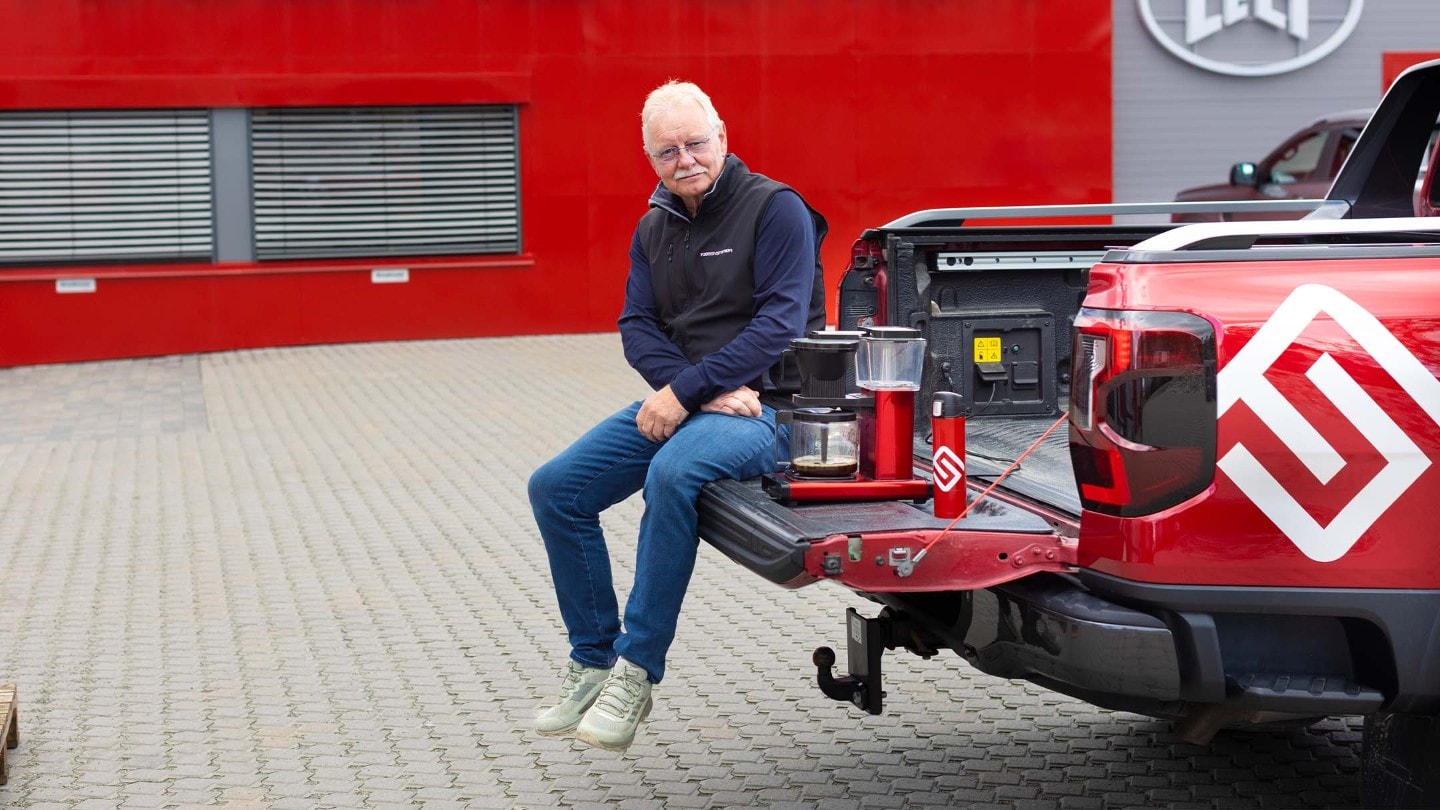 En mann sitter på bakluken til en rød Ford Ranger, med en kaffetrakter og kopp på lasteplanet.