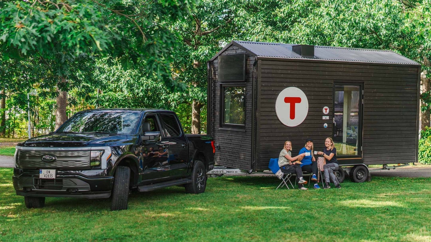 F-150 Lightning parkert ved den portable DNT-turisthytten og tre kvinner sitter utenfor og drikker kaffe.