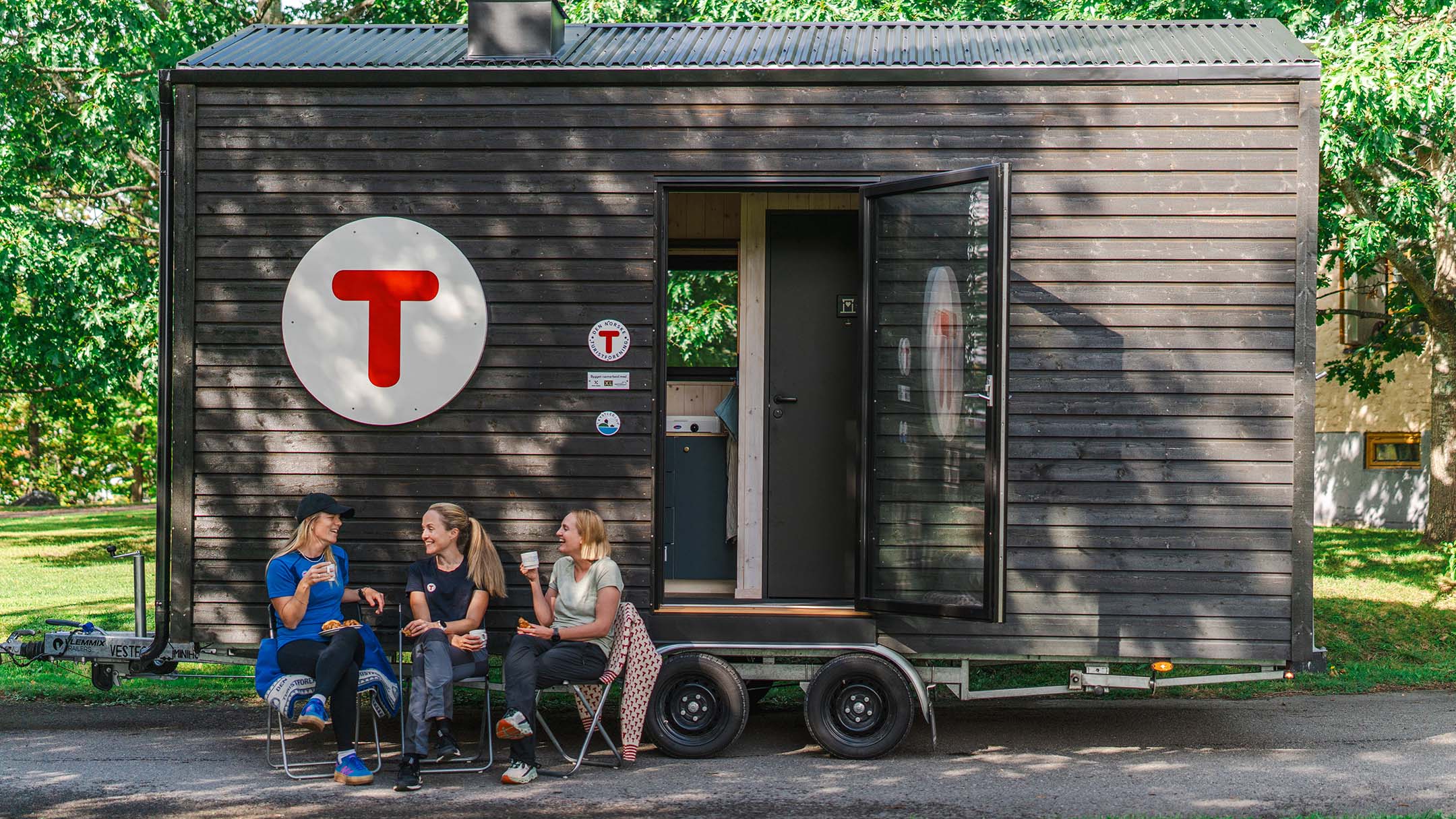 Tre kvinner sitter på stoler foran DNT-hytten og prater sammen.