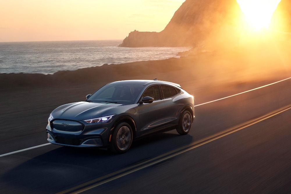 A Ford Mach-E drives on a seaside road at sunset