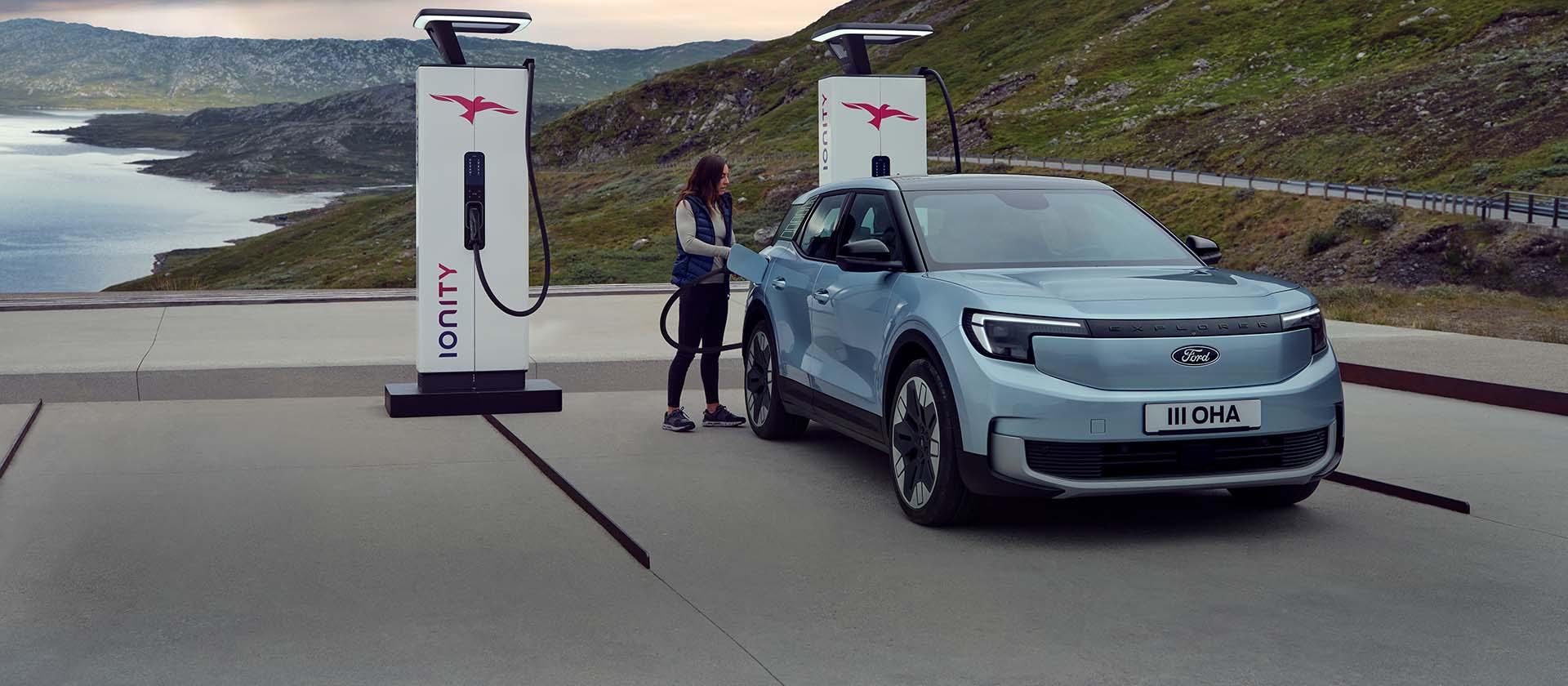 En kvinne kobler en blå, helelektrisk Ford Explorer til en offentlig ladestasjon med en kabel. Bilen er parkert ved fjell og vann.
