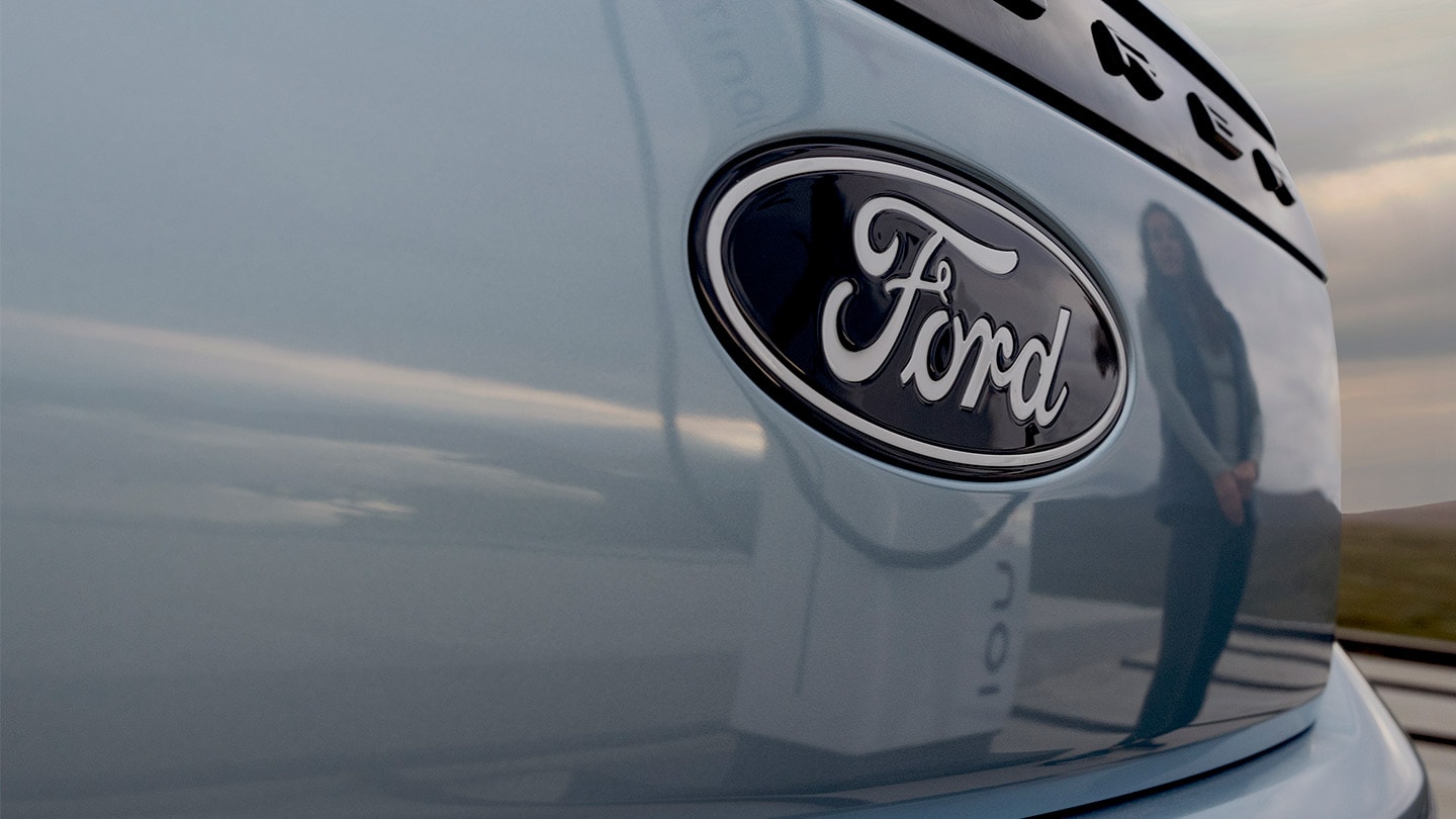  Ford-logo på en blå helelektrisk Ford Explorer med en offentlig ladestasjon reflektert i bilen.