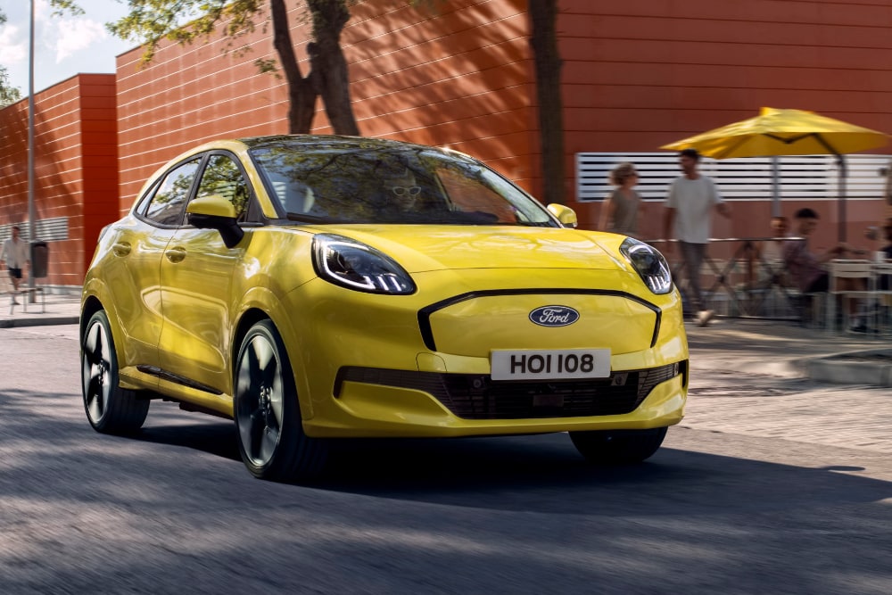 En gul Ford Puma kjører på en bygate med mennesker i bakgrunnen, sett fra front i trekvartsvinkel.