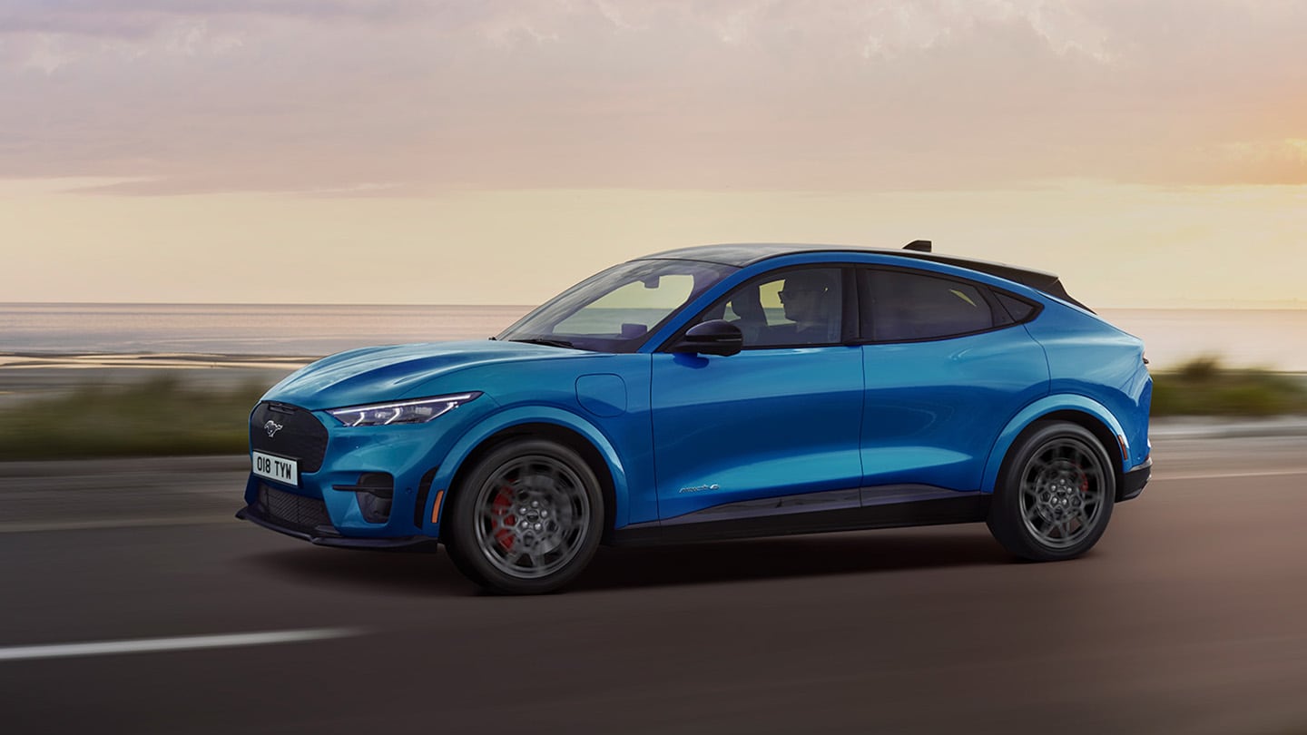 En blå, elektrisk Ford Mustang Mach-E vist fra siden mens den kjører langs en kystvei i skumringen.