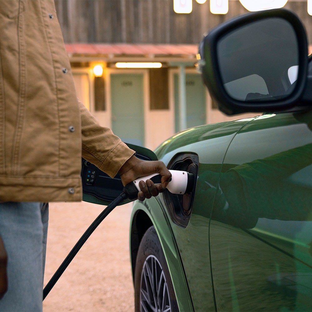 En person i en kremfarget jakke som plugger en elektrisk ladekabel inn i vedkommendes grønne, elektriske Ford Mustang Mach-E.