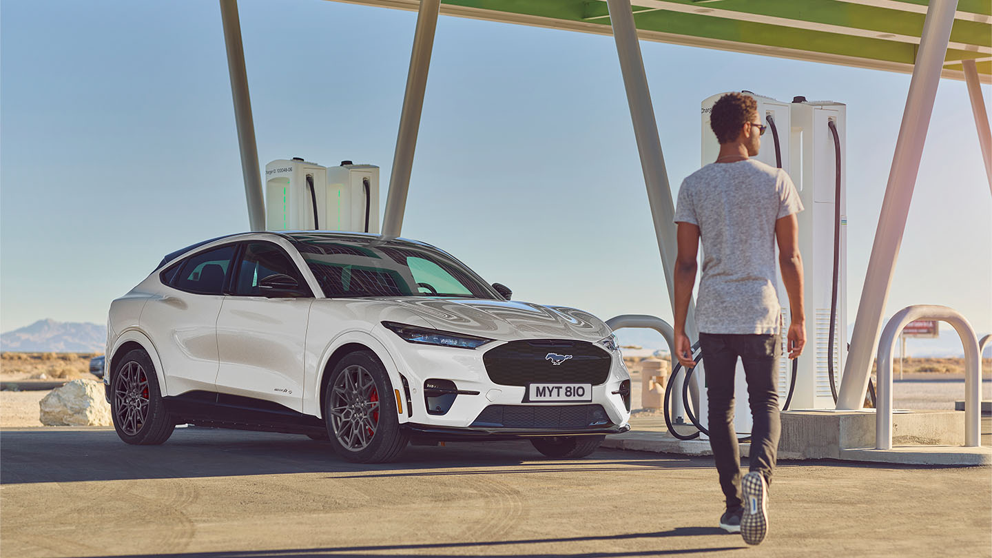 En hvit Ford Mustang Mach-E parkert utendørs utenfor en offentlig ladestasjon. En mann i en hvit T-skjorte går mot bilen.