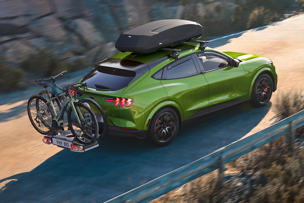 Rear view of a green Ford Mustang Mach-E with a roof box and a bike rack driving along a scenic road.