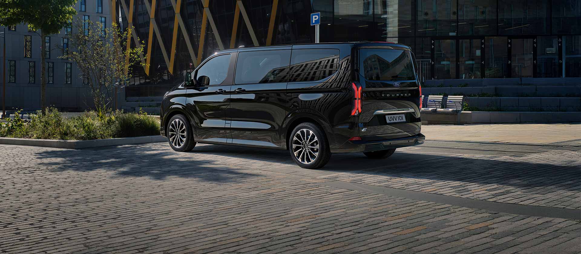 En svart Ford E-Tourneo Custom står parkert og viser siden og baksiden foran en moderne bygning.