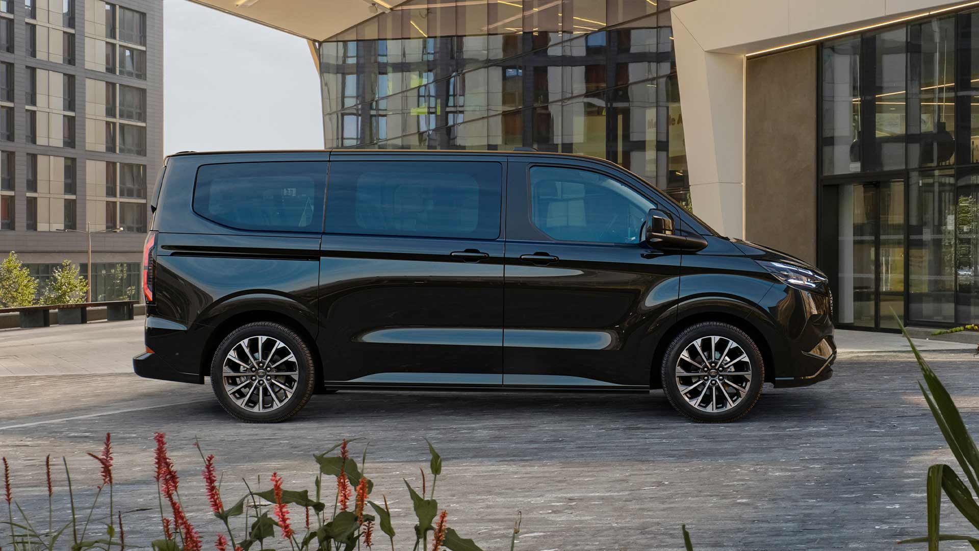 En svart Ford E-Tourneo Custom står parkert foran en moderne bygning.