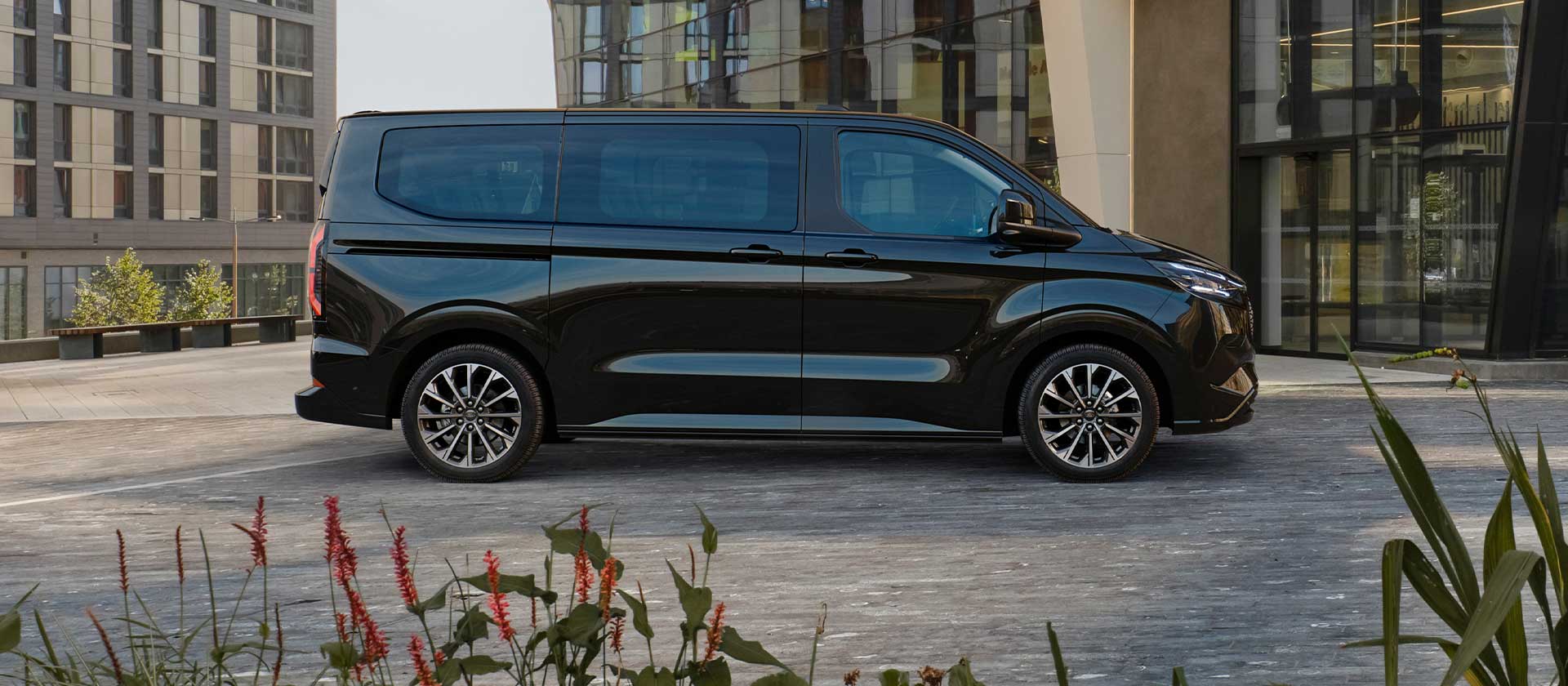 En svart Ford E-Tourneo Custom står parkert foran en moderne bygning.