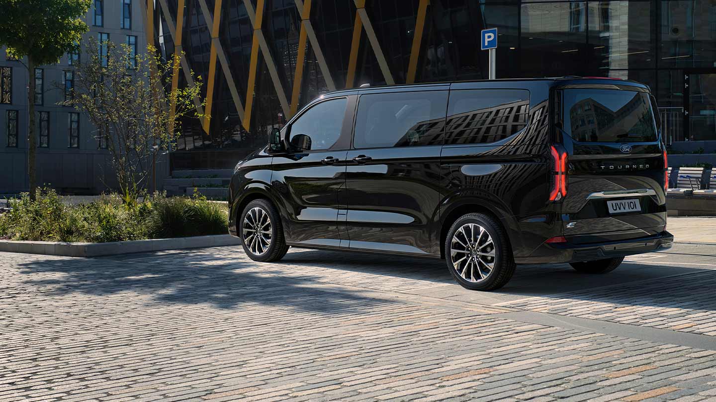 En svart Ford E-Tourneo Custom står parkert og viser siden og baksiden foran en moderne bygning.