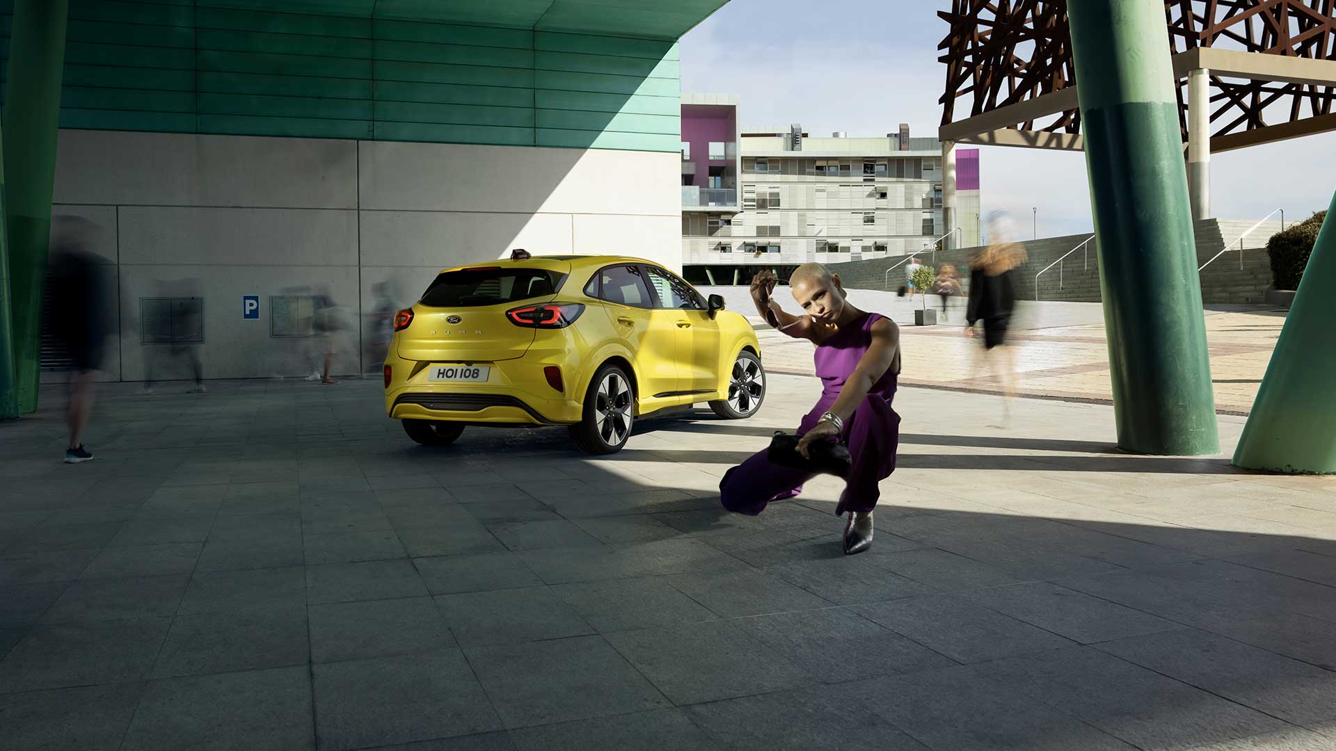 En gul elektrisk Ford Puma® vist bakfra, parkert under en bro ved siden av en person kledd i lilla.