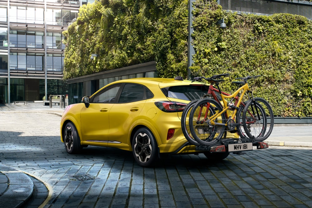 Gul helelektrisk Ford Puma parkert i et utbant miljø, med to sykler på et hengerfestemontert sykkelsativ. 