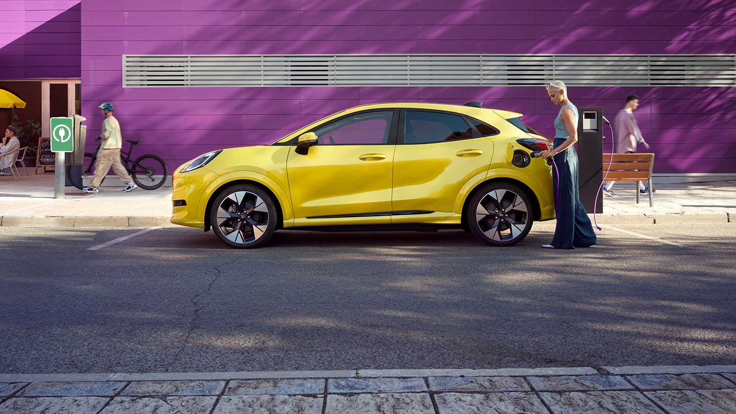 Sidevisning av en gul elektrisk Ford Puma® som er plugget inn i en offentlig ladestasjon mens den står på en parkeringsplass.