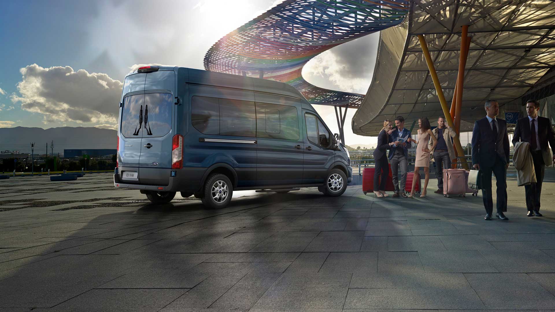 Sett skrått bakfra av en blå Ford Transit Minibuss parkert på en flyplass, med mennesker og bagasje i nærheten.