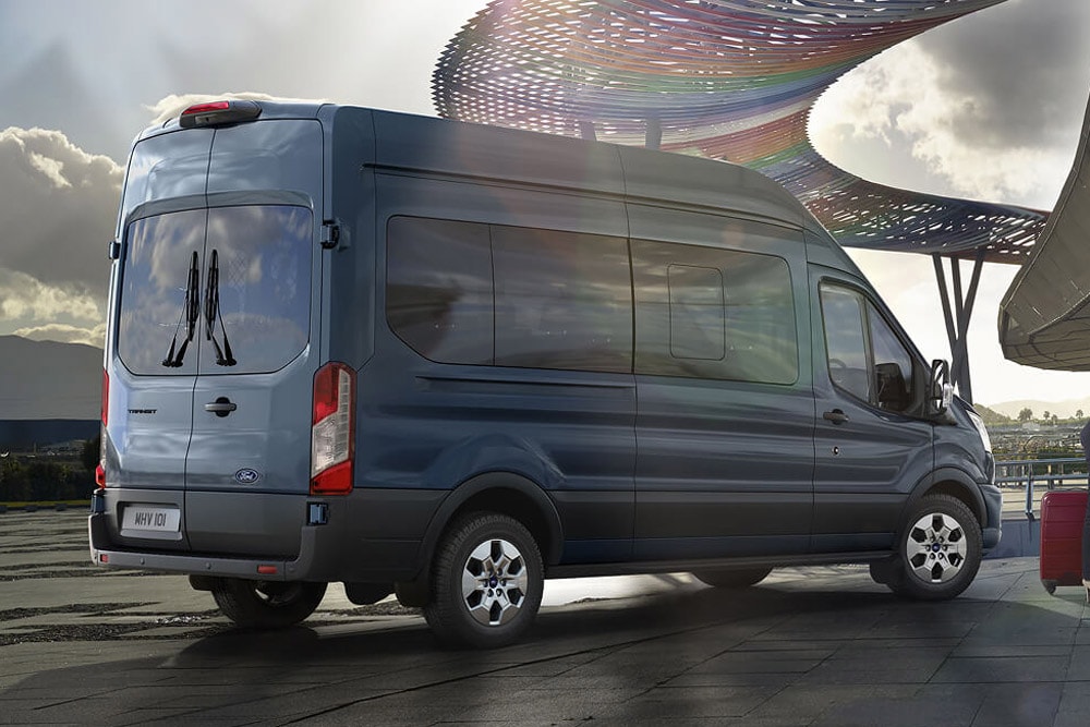 Sett skrått bakfra av en blå Ford Transit Minibuss parkert utendørs, med en moderne, fargerik konstruksjon i bakgrunnen.