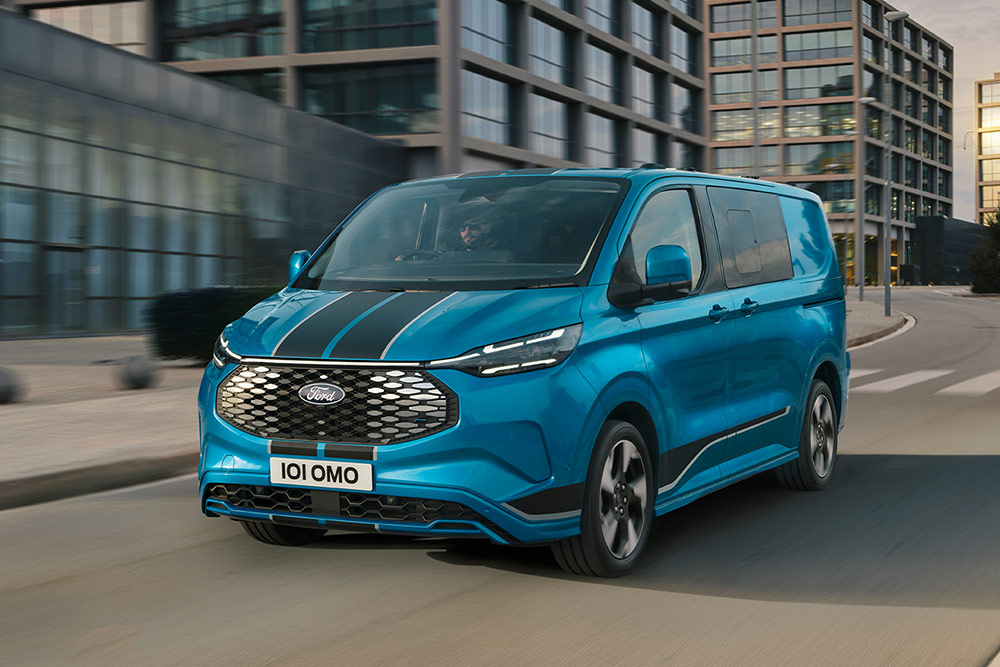 Blå Ford E-Transit Custom Sport dobbelkabin varebil som kjører på en bygate, ikke tilgjengelig i Norge.