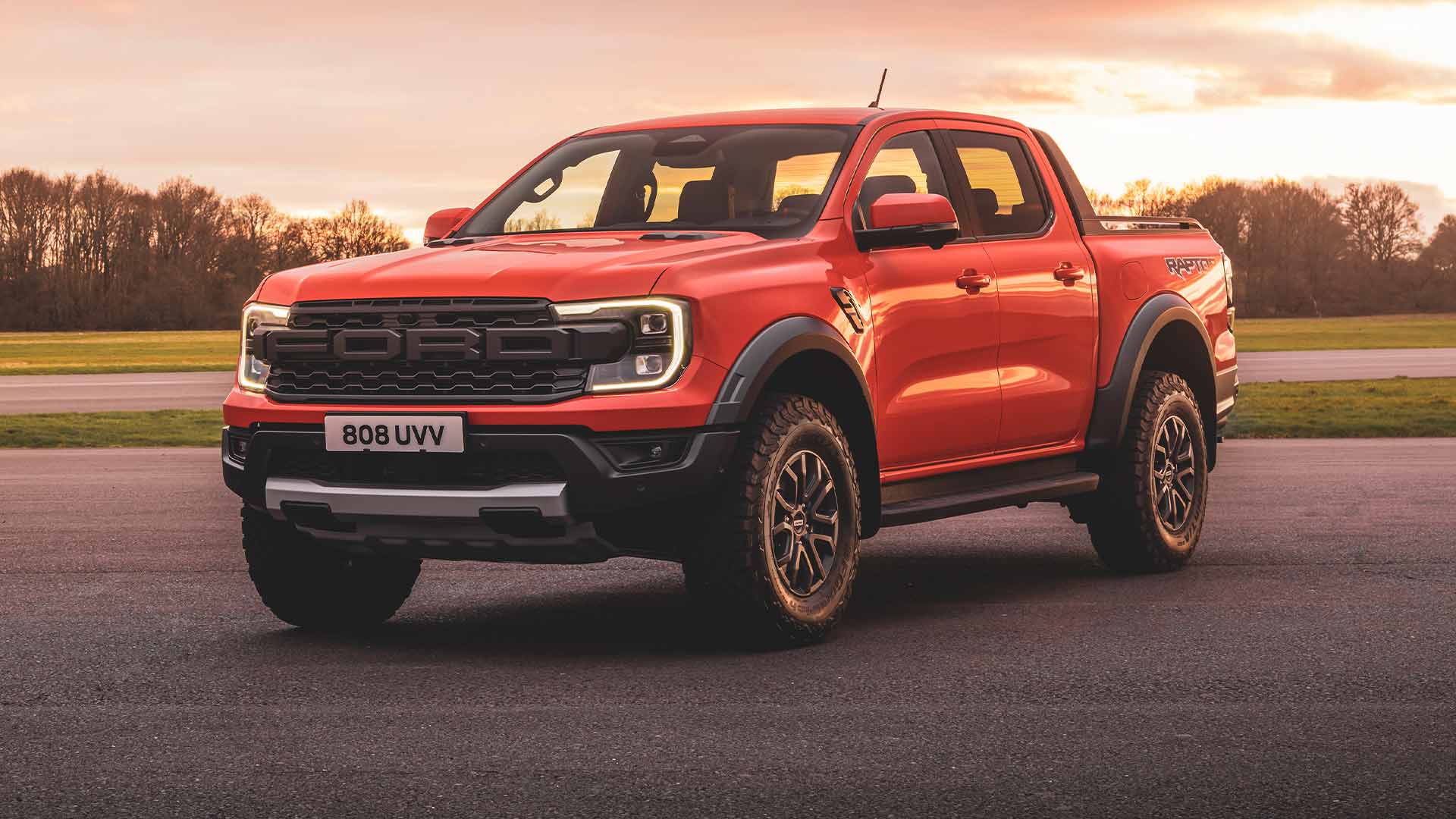   Ford Ranger Raptor i fargen Code orange parkert på an bane med frontlykter på.