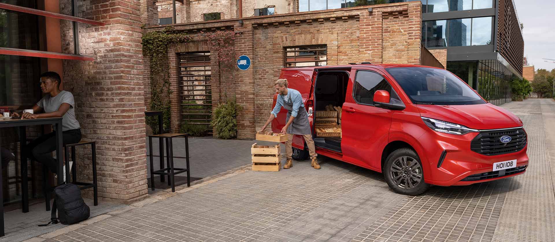 En person losser kasser av en rød Ford Transit Custom-varebil parkert foran en kaffebar.