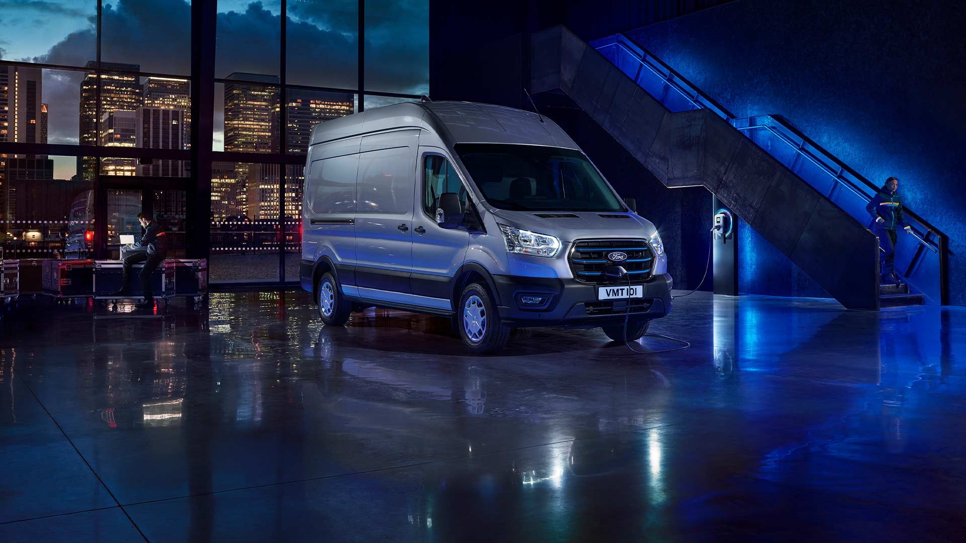 Sølvfarget Ford E-Transit elektrisk varebil parkert innendørs med en bysilhuett synlig gjennom store vinduer.