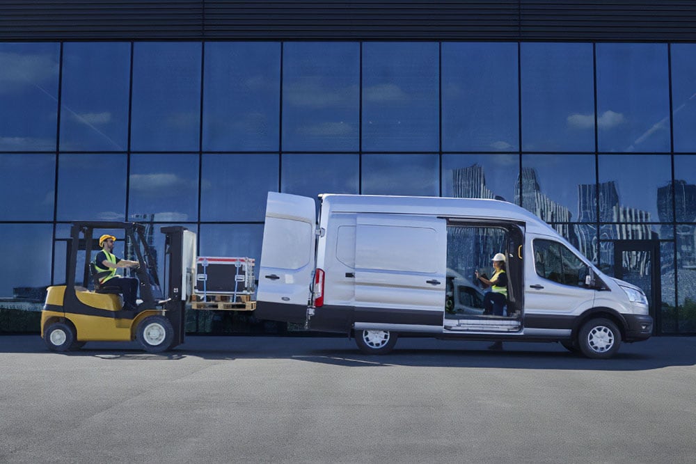 To personer laster last inn i en sølvfarget Ford Transit-varebil med gaffeltruck.