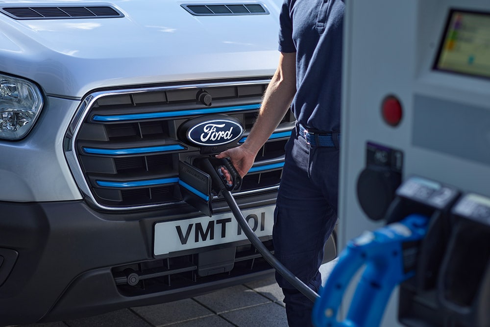 Nærbilde av en person som kobler en ladekabel til en sølvfarget Ford E-Transit elektrisk varebil.