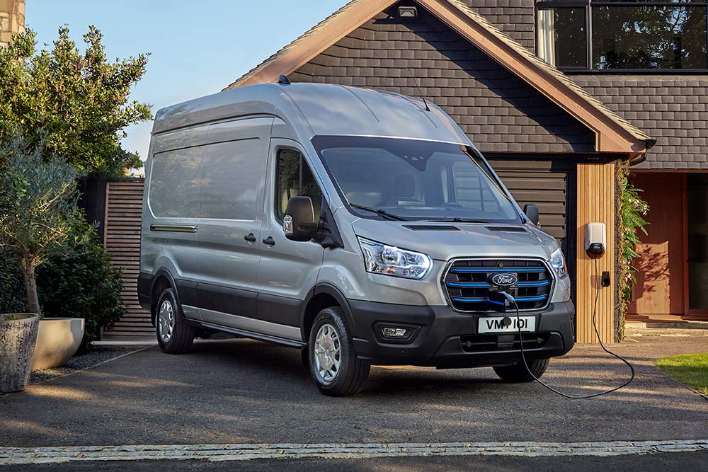 Sølvfarget Ford E-Transit varebil parkert i en oppkjørsel ved siden av et hus, koblet til en ladestasjon.