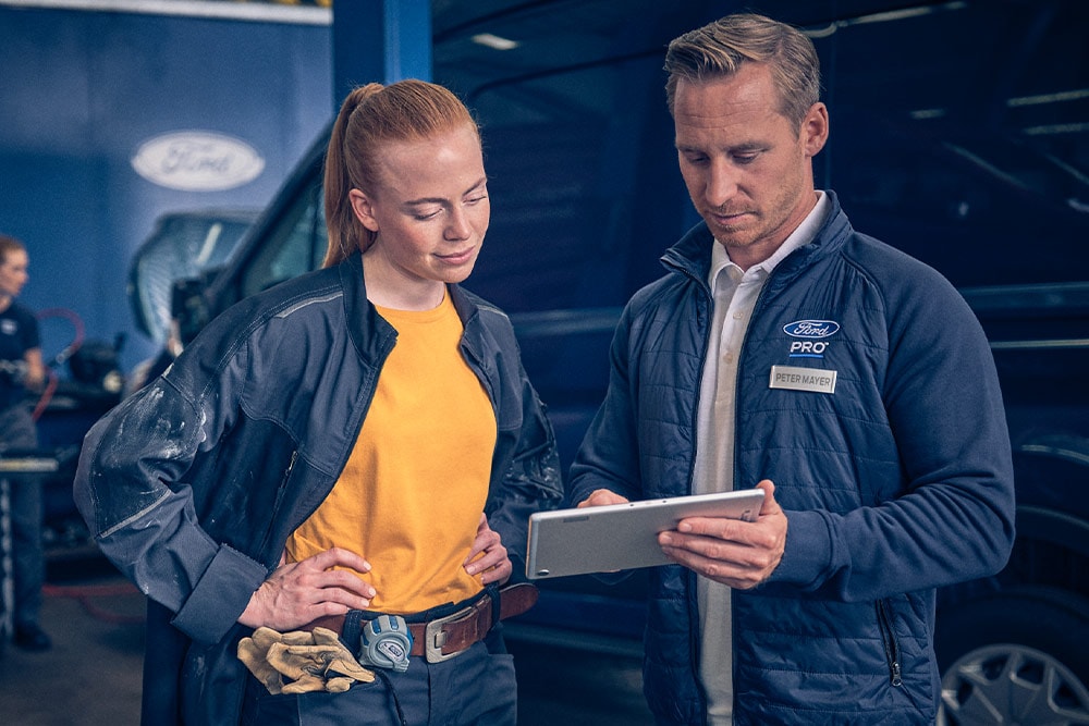 To mekanikere i en garasje som ser på et nettbrett foran en Ford E-Transit elektrisk varebil.