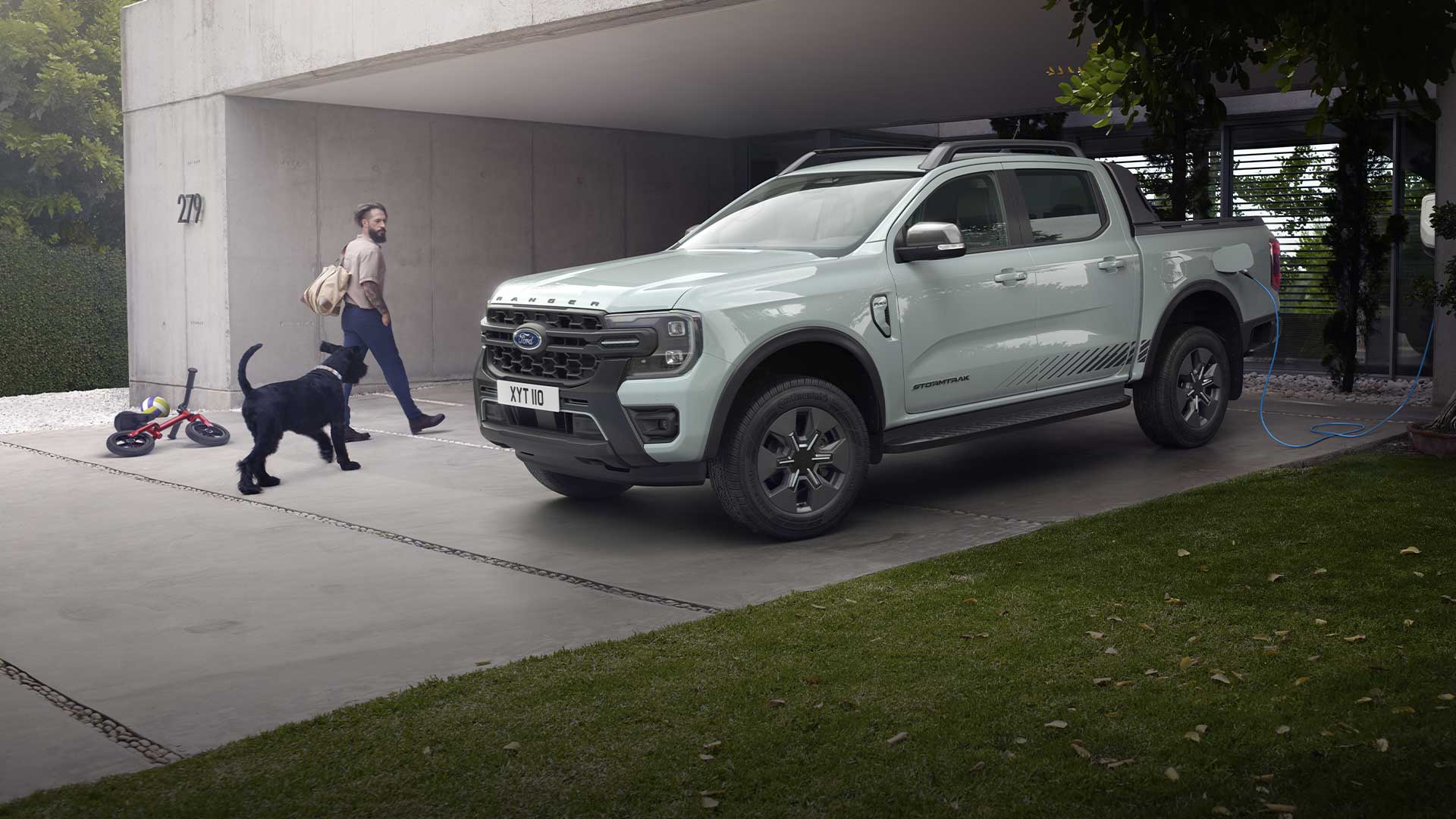 En mann og en hund går mot en grå, ladbar lett Ford Ranger-hybridlastebil som lades i en oppkjørsel.