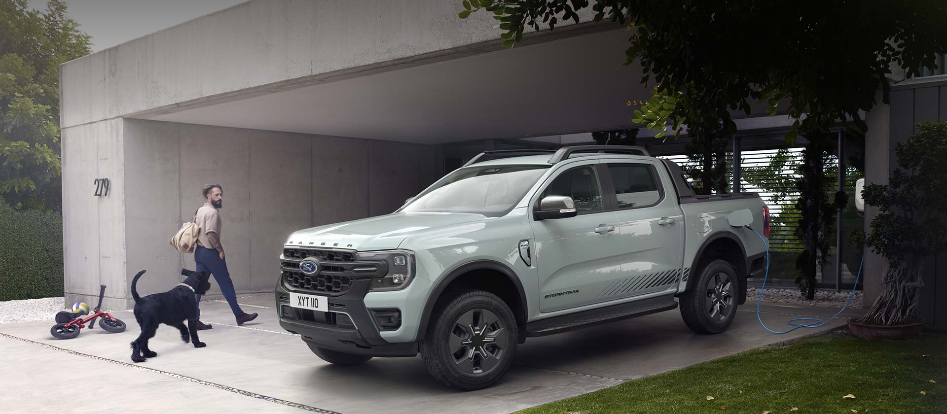 En mann og hunden hans går inn i et moderne hus idet de passerer en Ford Ranger Stormtrak PHEV som står til lading.