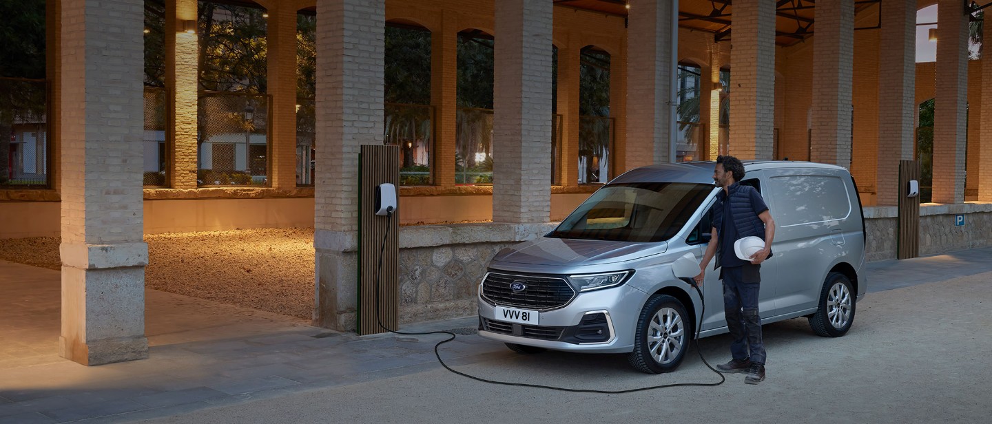 En mann som holder en vernehjelm plugger en ladekabel fra veggboks inn i splitter nye Ford Transit Connect i Stardust Silver.