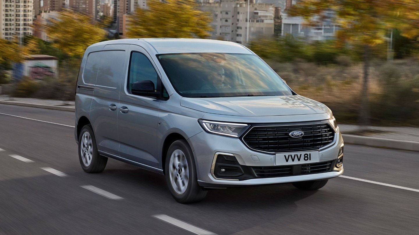 Visning av splitter nye Ford Transit Connect Limited i Stardust Silver som kjører på en vei, med kromdetaljer på eksteriøret og automatiske frontlykter. 