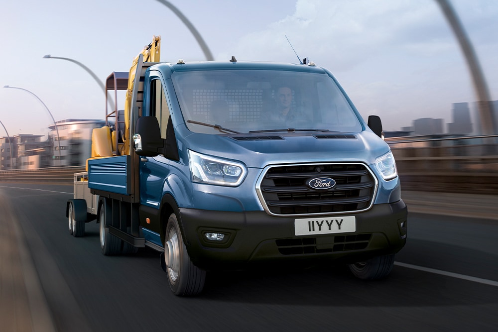 Visning av fra frontgrillen til Ford Transit Chassis Cab som frakter en gravemaskin mens den kjører langs en byvei. 