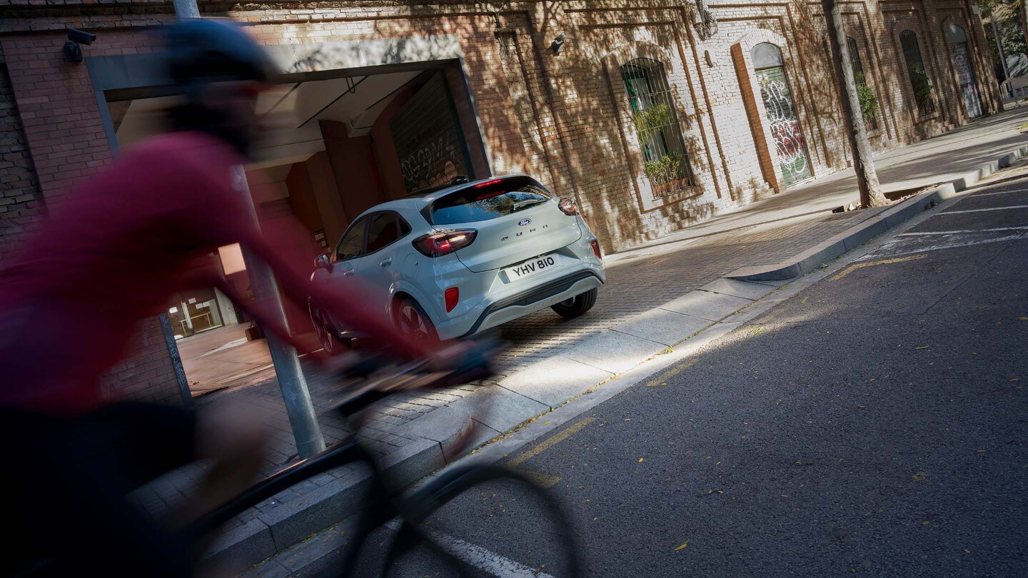Grå Ford Puma rygger ut av oppkjørselen, syklist i rødt passerer.