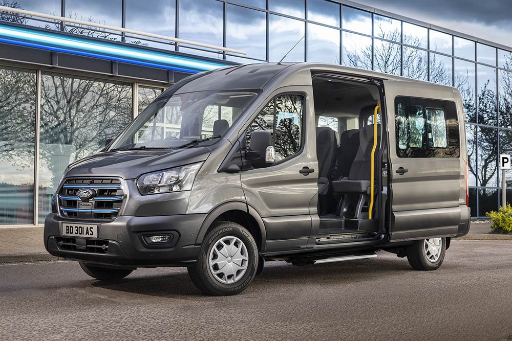 Sidevisning av grå Ford Pro Transit med åpne dører, parkert foran en moderne bygning.