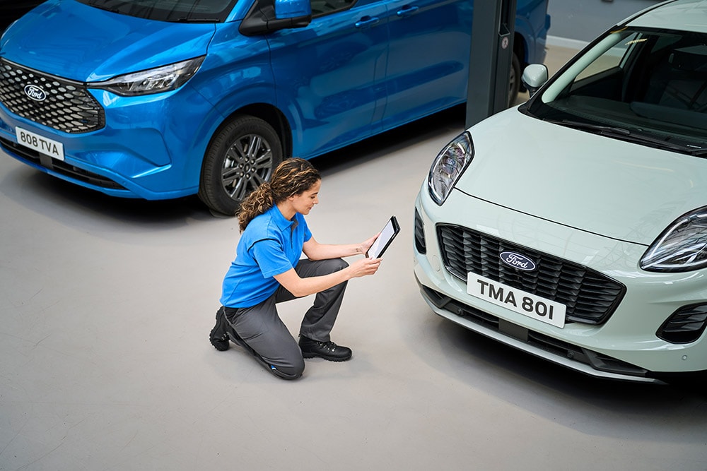 En tekniker kneler mellom en blå Ford-varebil og en hvit Ford Puma, og ser på et nettbrett.