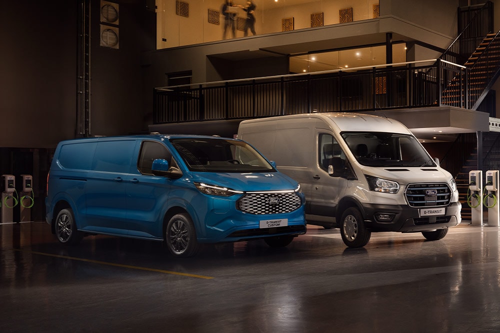 En blå Ford E-Transit Custom og en sølvfarget Ford E-Transit som lader på en parkeringsplass.