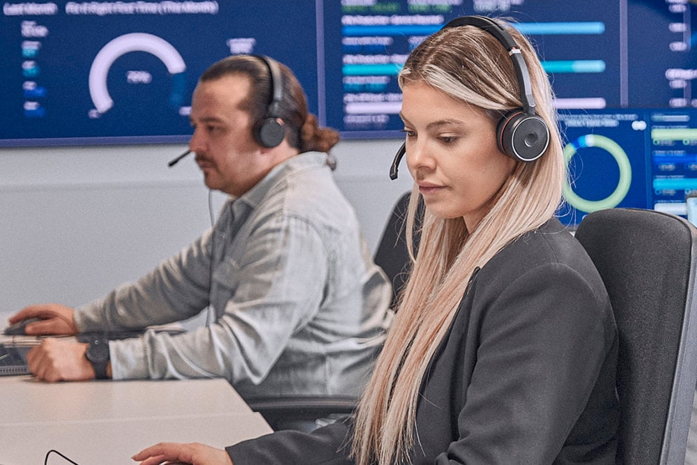 To personer med hodesett jobber ved datamaskiner i et moderne kontrollrom.