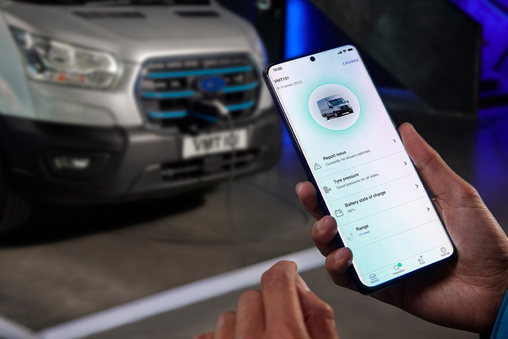 En person holder en smarttelefon som viser en Ford-app, med en Ford E-Transit Custom som lader i bakgrunnen.