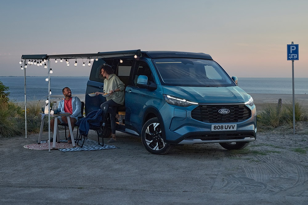 To bobiler slapper av ved en blå Ford Pro Transit Custom Nugget, et parkeringsområde ved stranden i bakgrunnen.