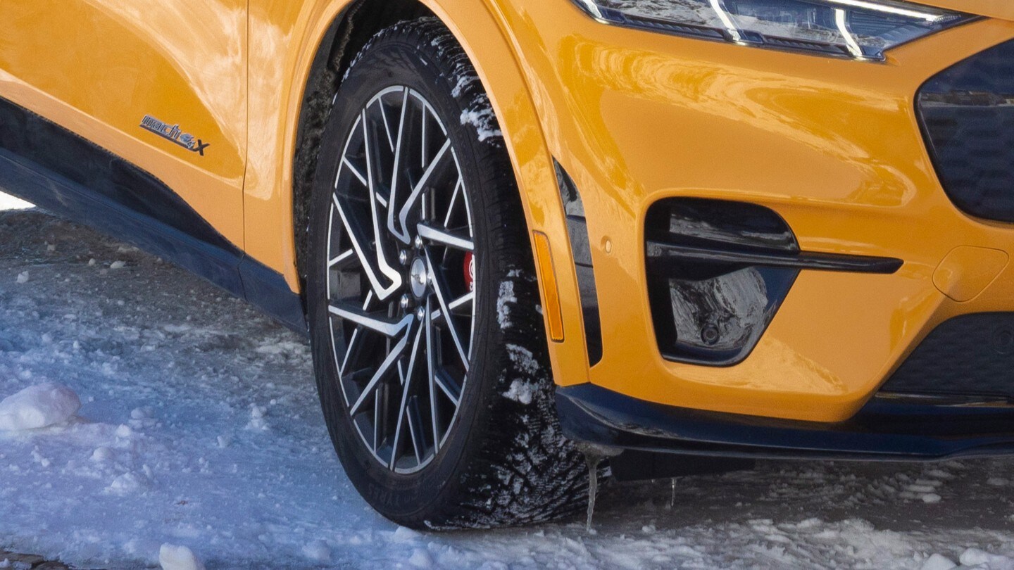 Et nærbilde av et forhjul og en frontfanger på en Cyber Orange Mustang Mach-E, parkert på et snødekket underlag. 