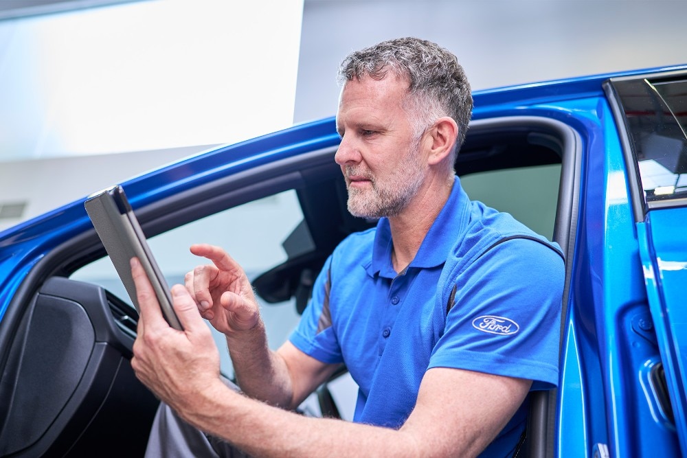 A Ford technician sits in a blue Ford vehicle, using a tablet to check vehicle systems.